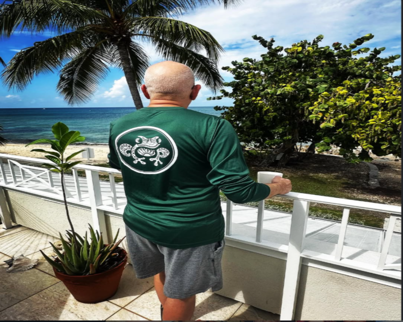 Man wearing a green shirt with a logo, standing on a balcony overlooking the ocean.