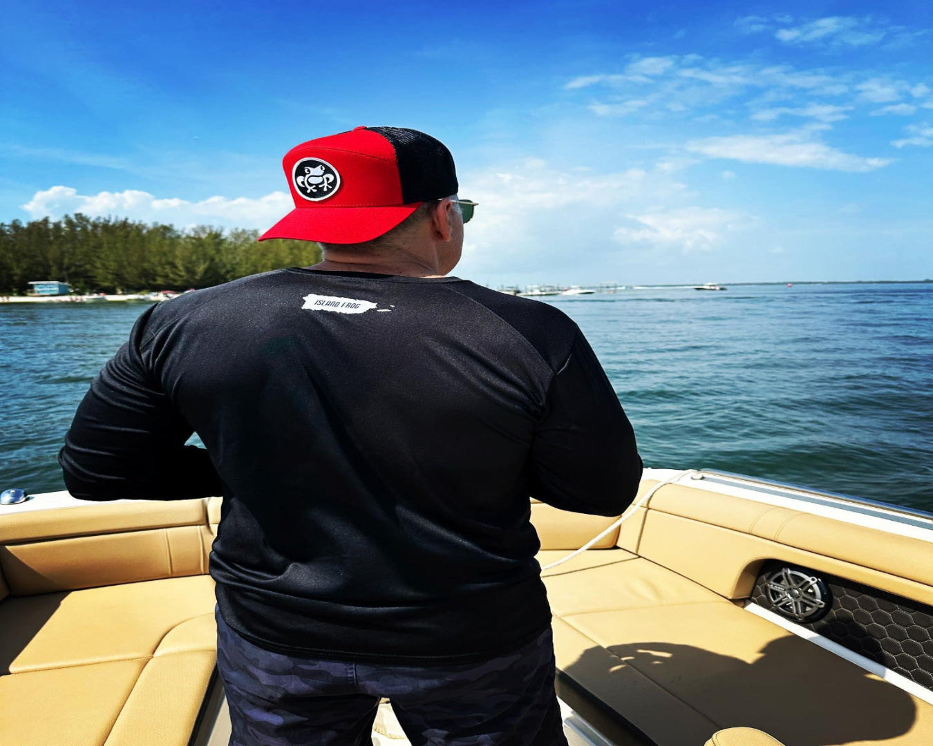 Man wearing a red and black cap and black shirt on a boat with a scenic view of water and sky.